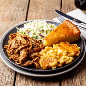 What Pairs Well with Pulled Chicken? 1 A plate of pulled chicken with coleslaw, mac and cheese, and cornbread on a rustic wooden table